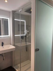 Modernes Badezimmer im Komfort-Doppelzimmer der Aal-Kate Wuppertal mit großer Dusche, Glasabtrennung, Regenduschkopf und Waschbecken mit Spiegel.
