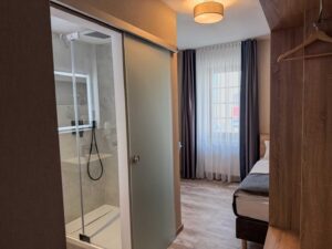 Einblick in das Komfort-Einzelzimmer der Aal-Kate Wuppertal mit Blick auf Dusche, Fenster mit Tageslicht und stilvollem Innenraum in Holzoptik.